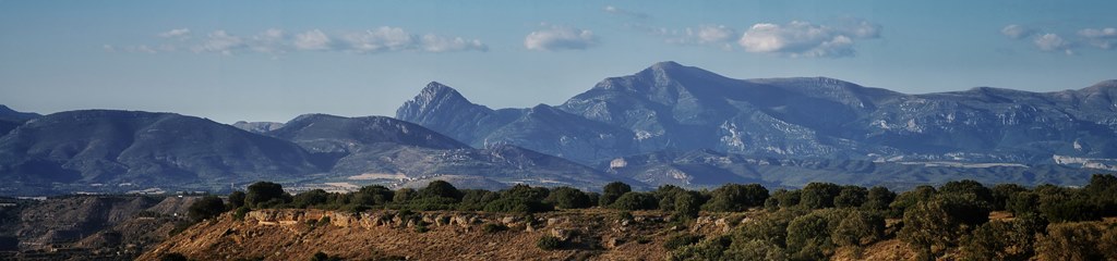 Fotografía panorámica de una cordillera nevada con árboles en primer plano y un cielo azul con nubes.